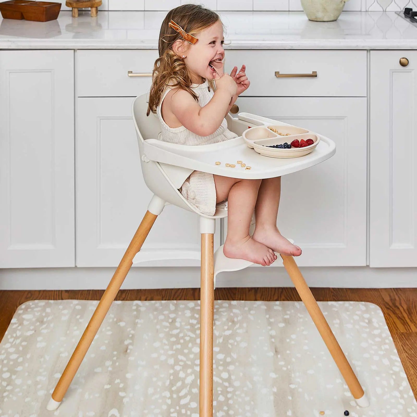 Fawn brown and white animal print high chair mat shown in a kitchen under a highchair with toddler girl sitting in the chair smiling and eating fruit