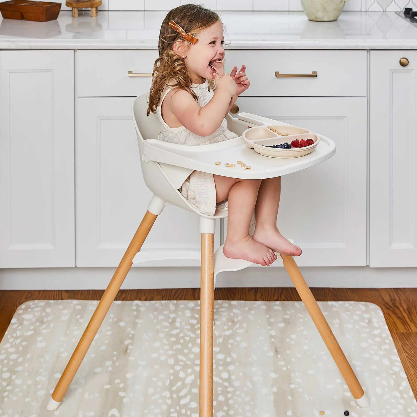 Fawn brown and white animal print high chair mat shown in a kitchen under a highchair with toddler girl sitting in the chair smiling and eating fruit