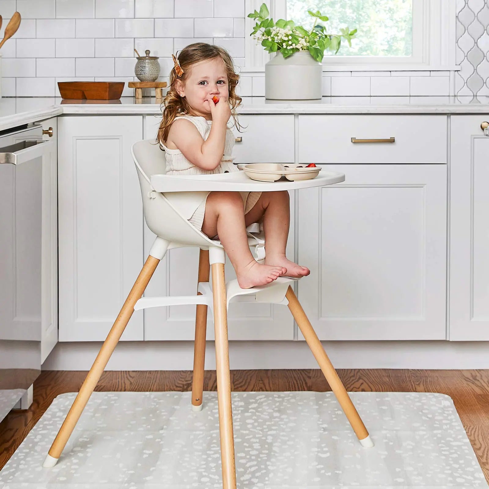 Fawn silver gray and white animal print high chair mat shown in a kitchen underneath a highchair with toddler girl sitting in the chair eating fruit