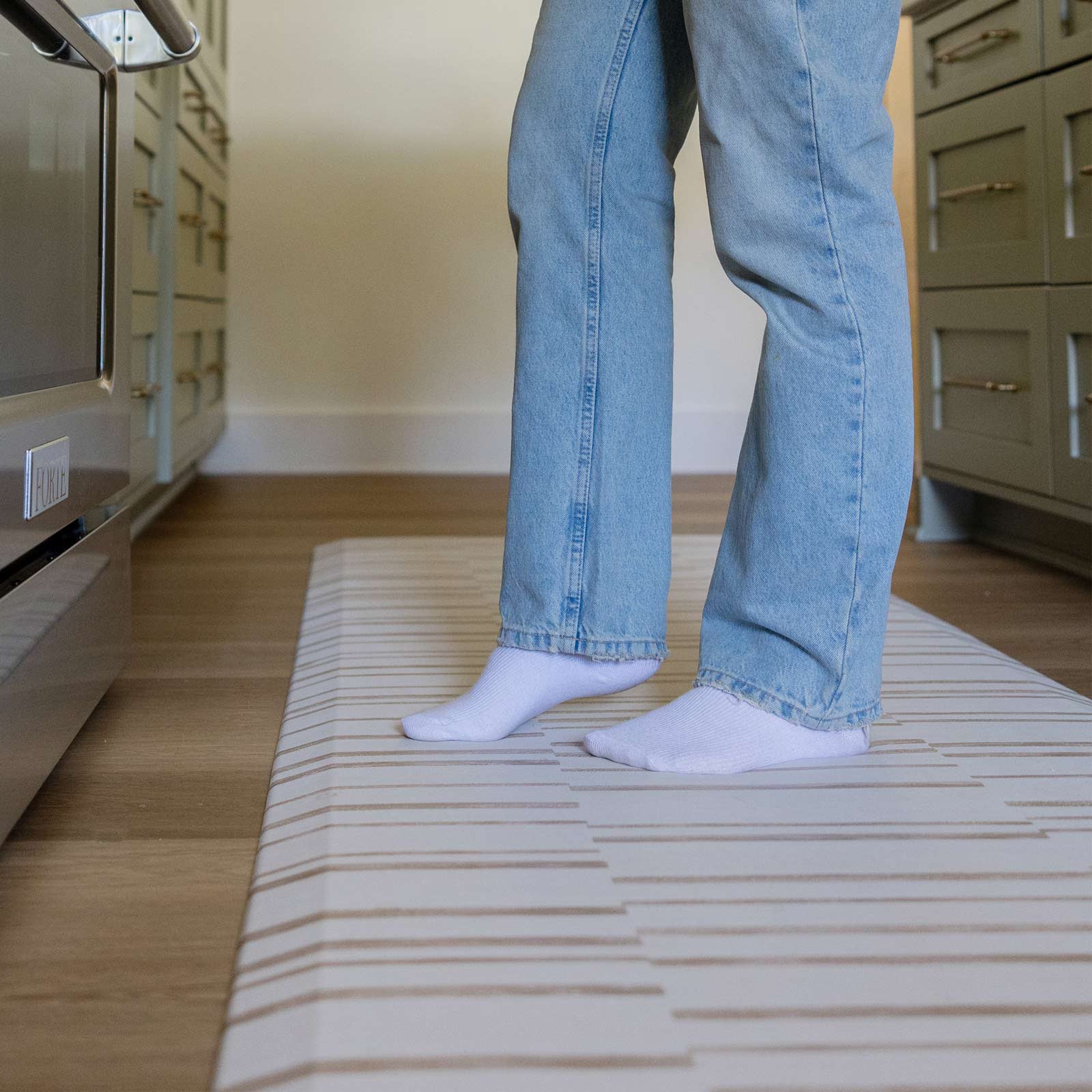 Nara natural beige and cream inverted stripe standing mat shown in kitchen with womans feet standing on the mat in front of stove. Shown in size 22x72