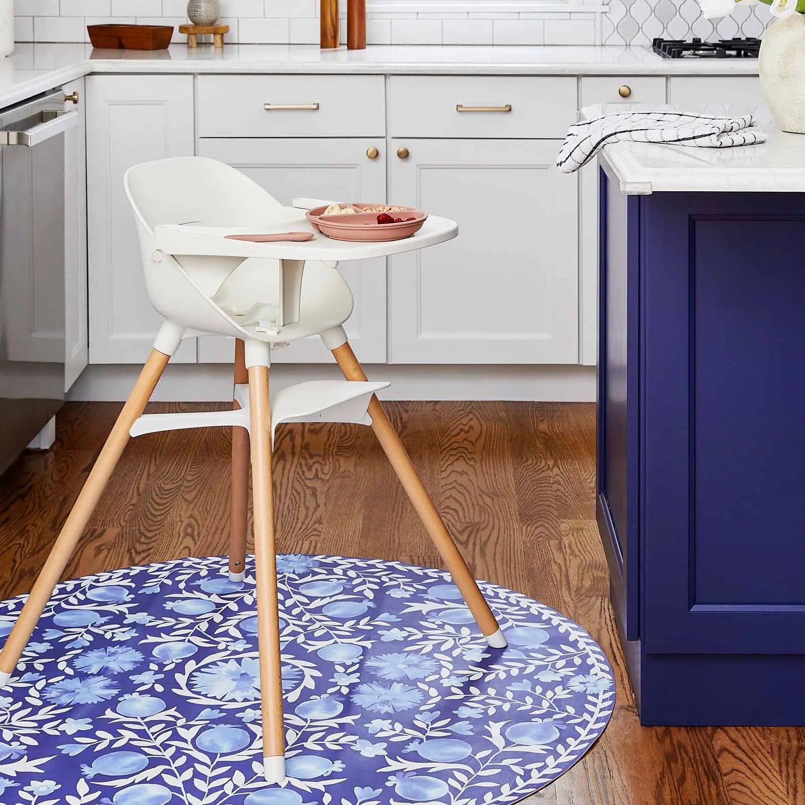 Suzette deep sea blue and white floral vine print highchair mat shown in kitchen under a highchair with a plate of fruit on it