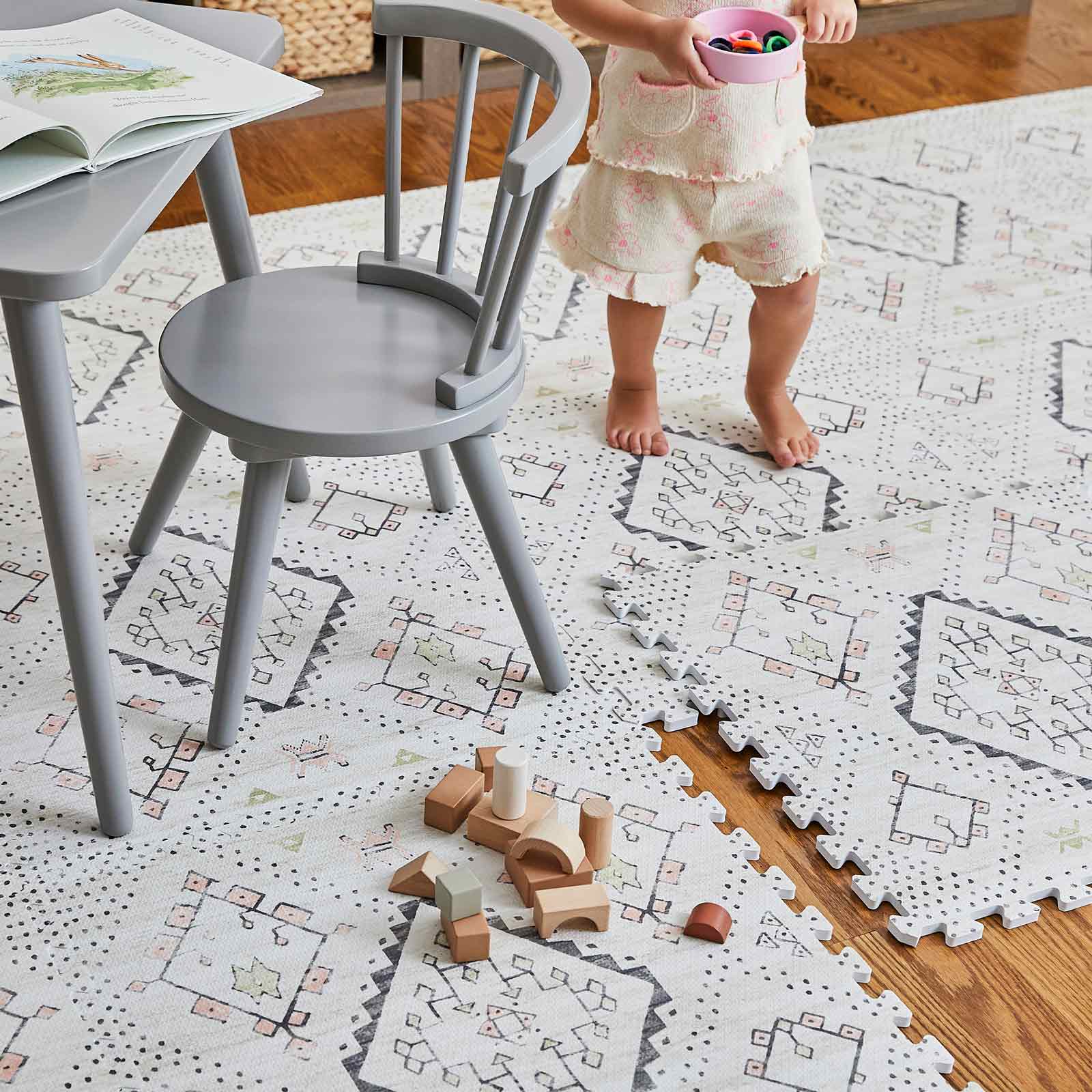 Child playing with toys on a patterned play mat in a room with a chair and table.
