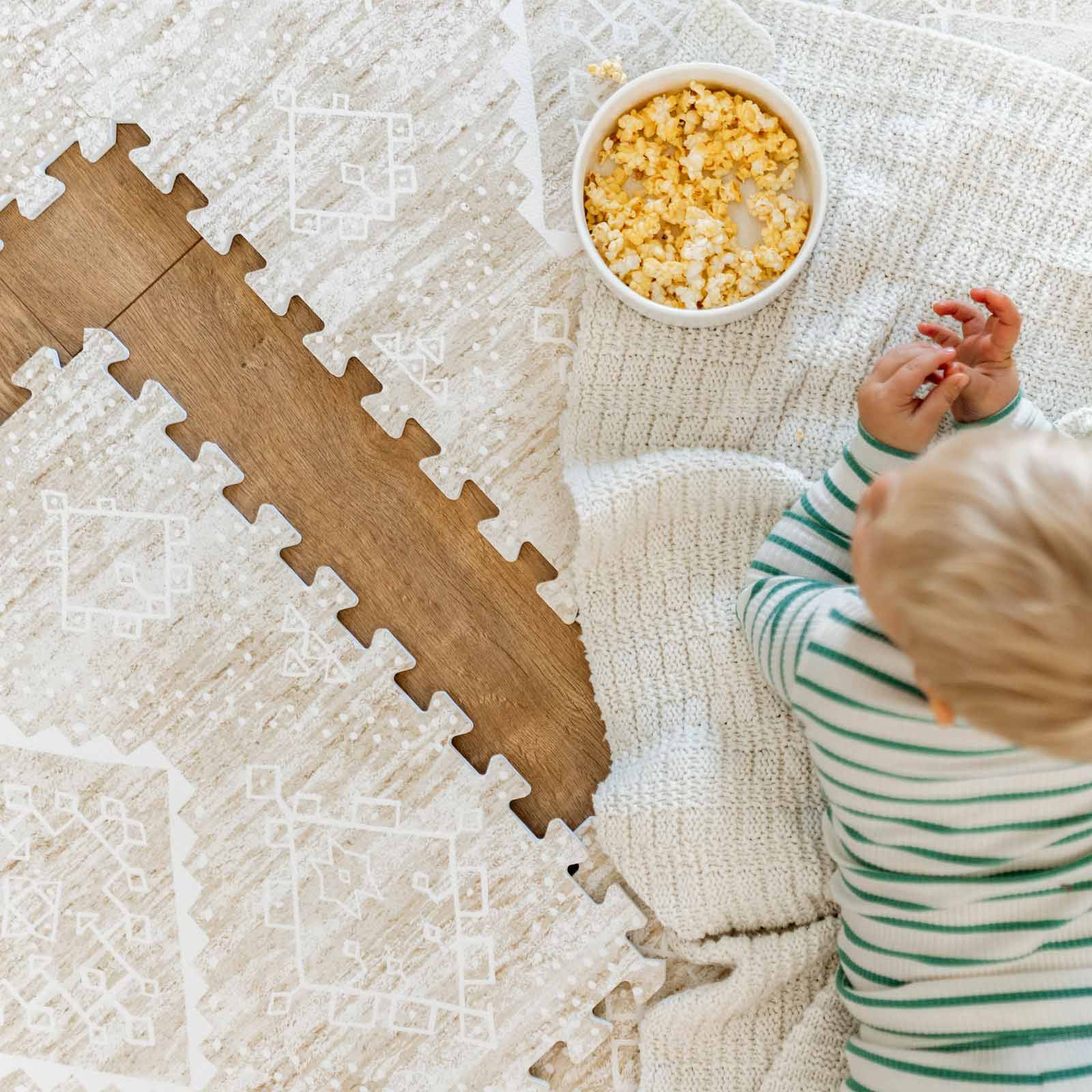 Ula straw neutral tan boho print play mat shown with 1 tile exposed and toddler laying on the mat eating popcorn