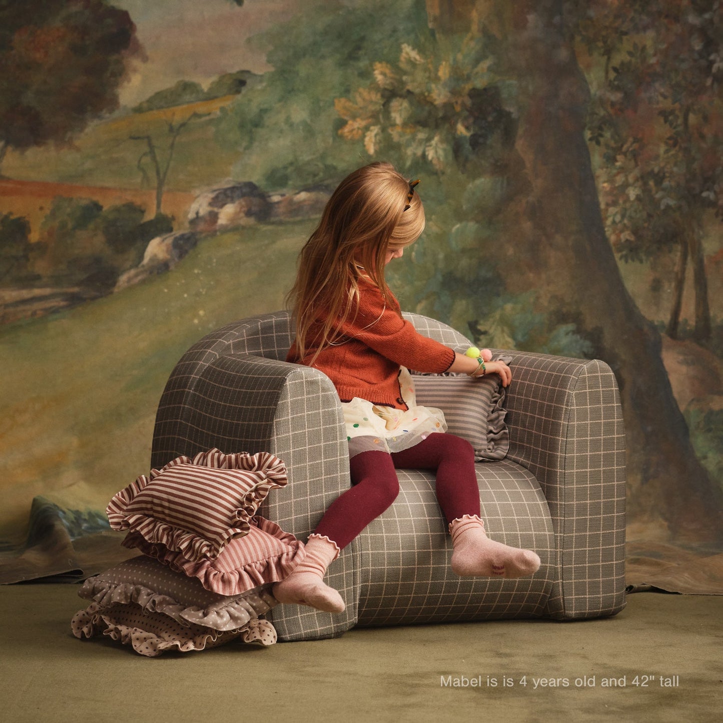 Young girl sitting on a checkered chair with pillows in a painted landscape setting