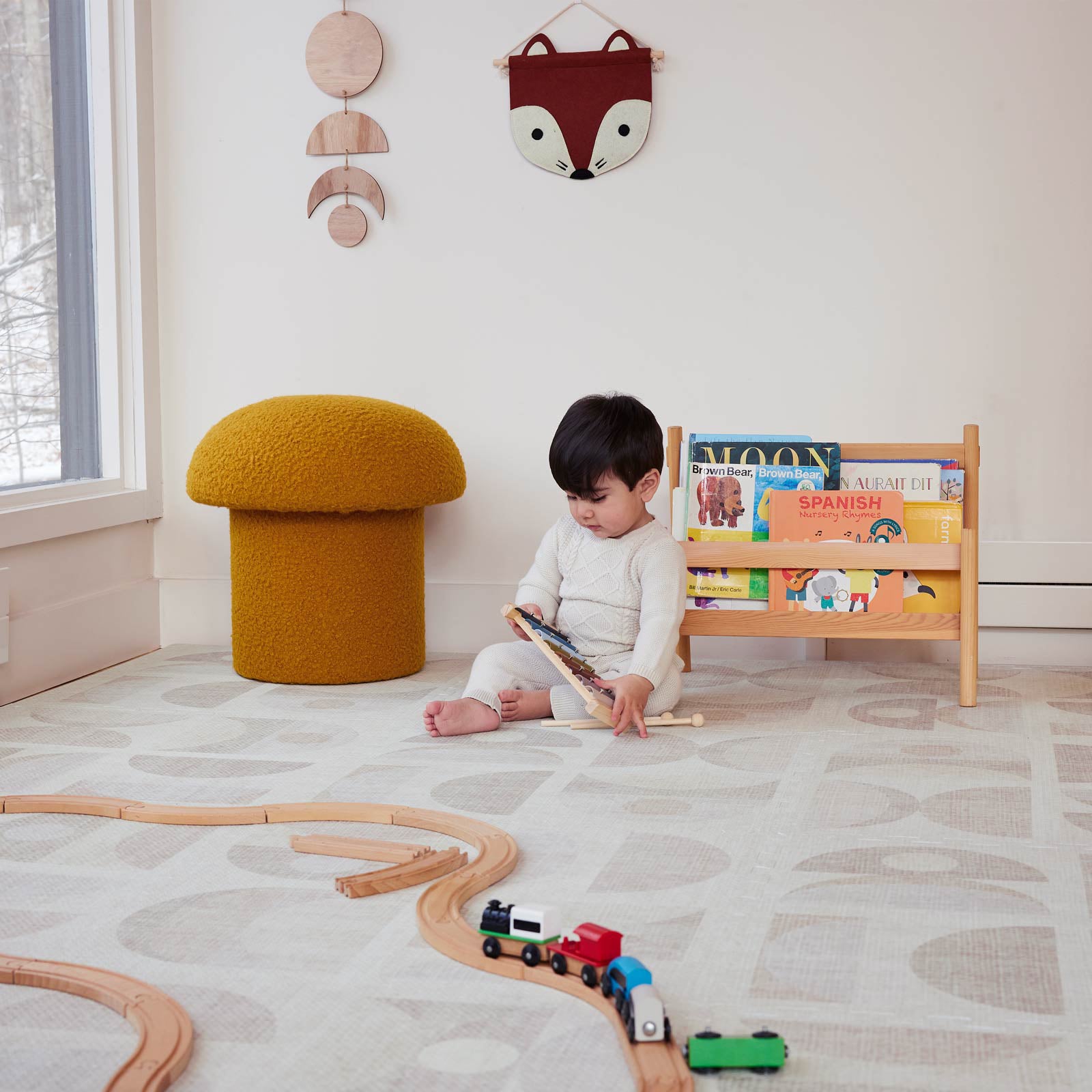 Luna sandstone neutral geometric print play mat shown in play room with wooden train track toys, mustard mushroom ottoman in the corner and baby boy sitting by a wooden bookstand opening a book