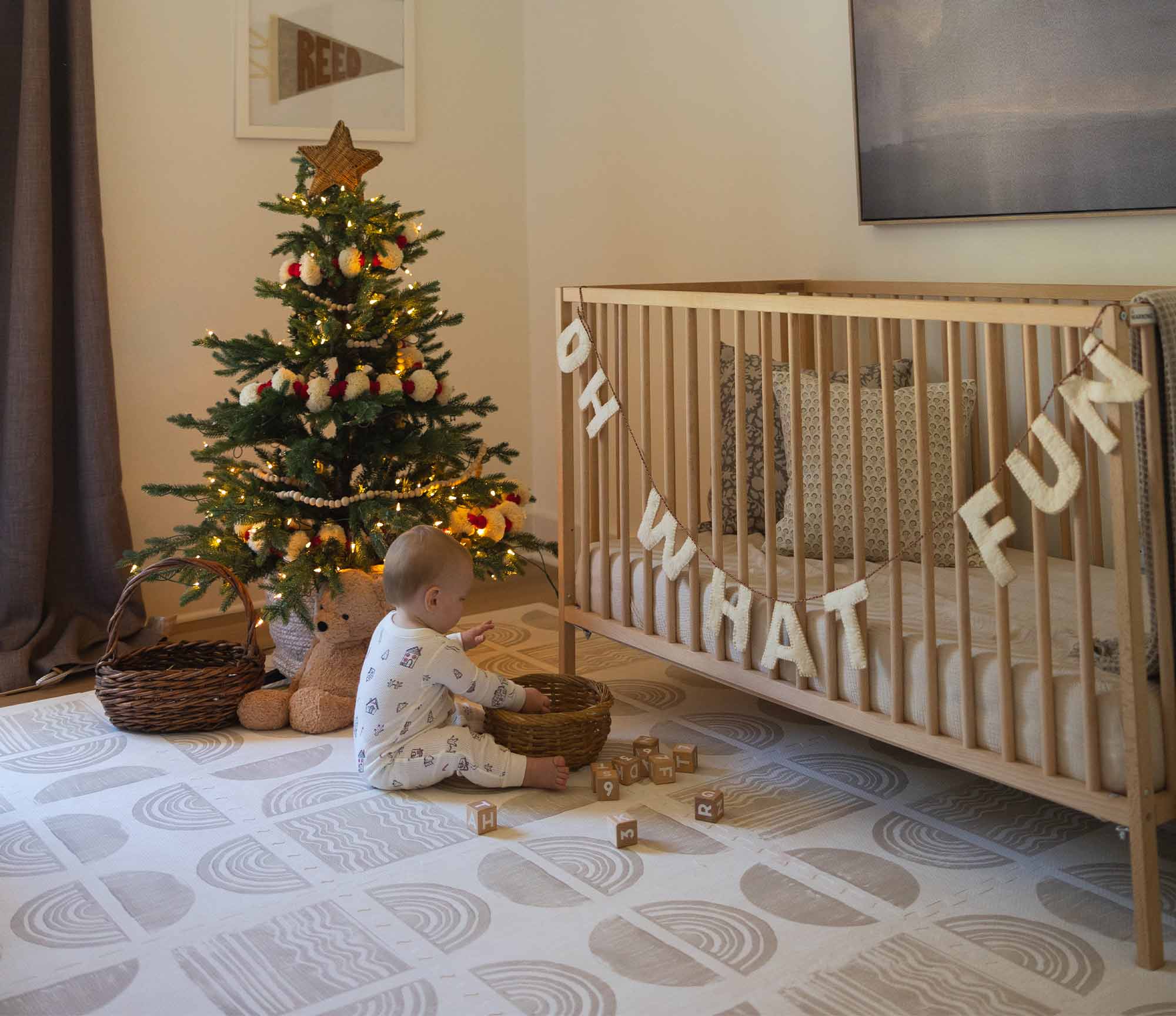 Ada Pebble white and tan geometric print play mat shown in a nursery with baby playing on the mat in front of a christmas tree and crib with an "oh what fun" banner strung across