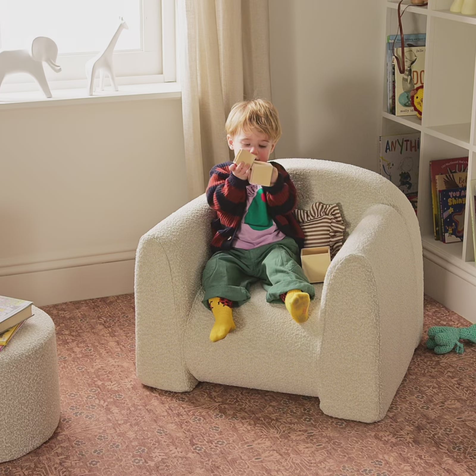 Child sitting in white boucle fabric play chair on top of a patterned play mat next to a bookshelf