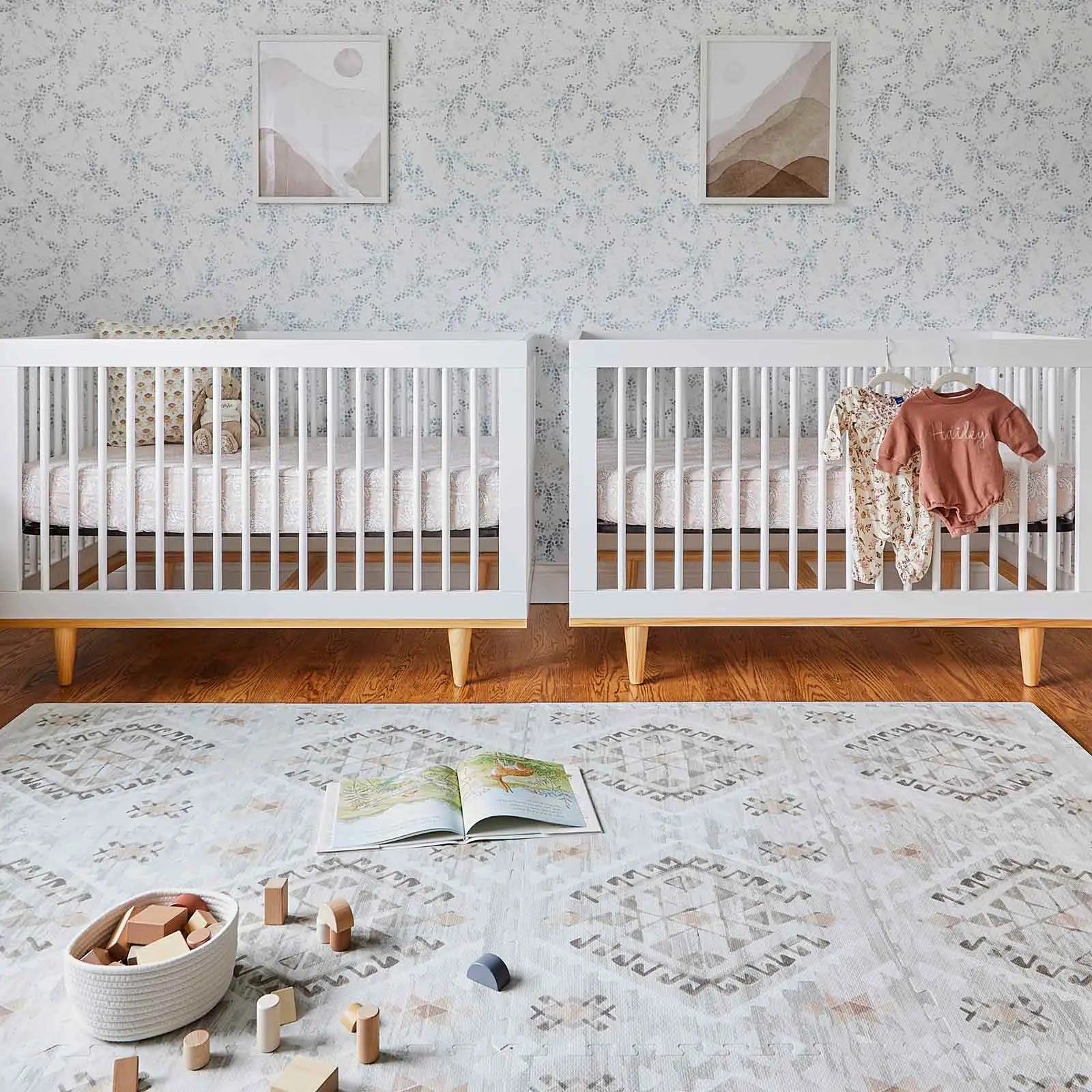 Nalla Basalt grey and sage green boho print play mat shown in nursery with 2 cribs with a book and blocks on the mat