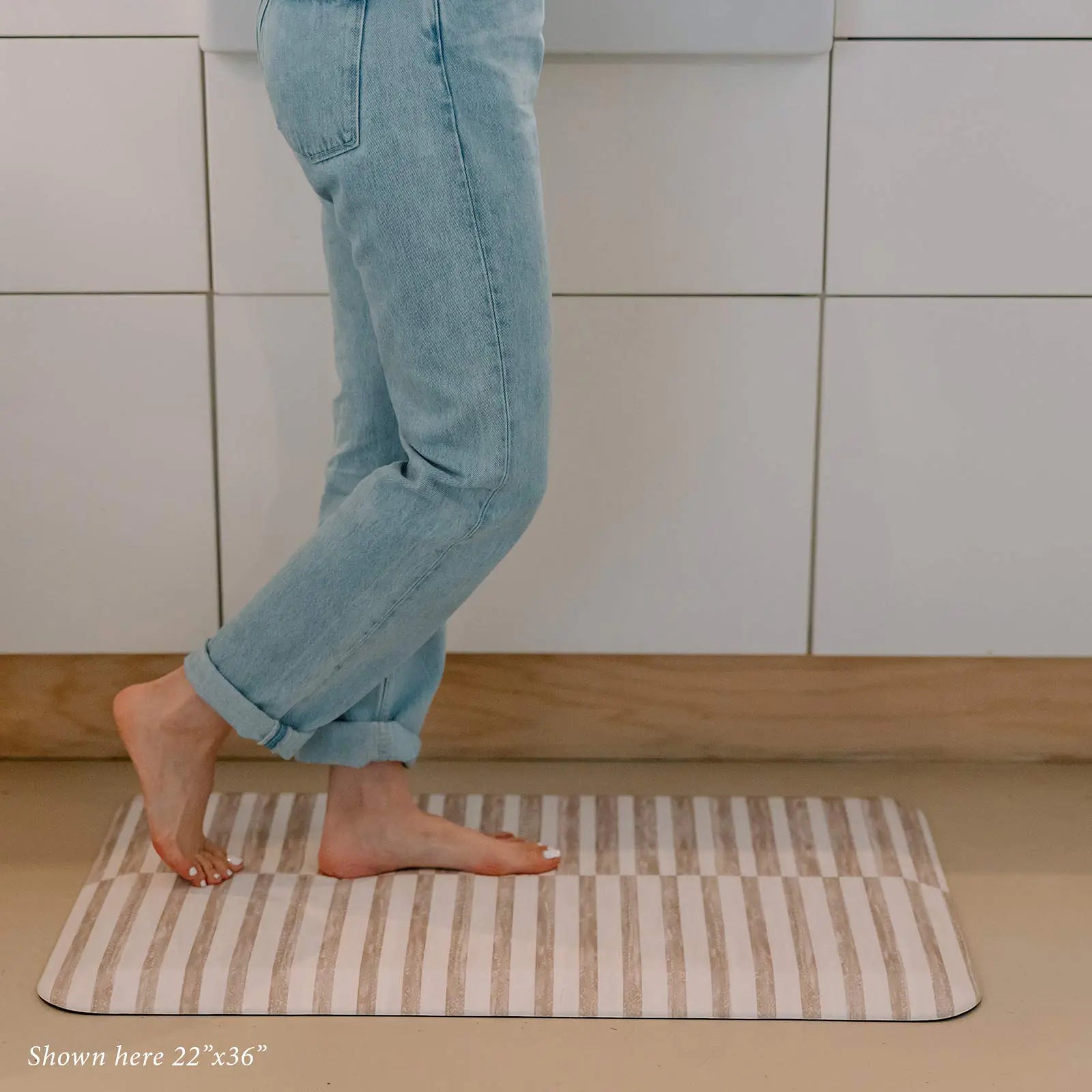 Reese chai beige and white inverted stripe standing mat shown in kitchen in size 22x36 with womans feet standing on the mat