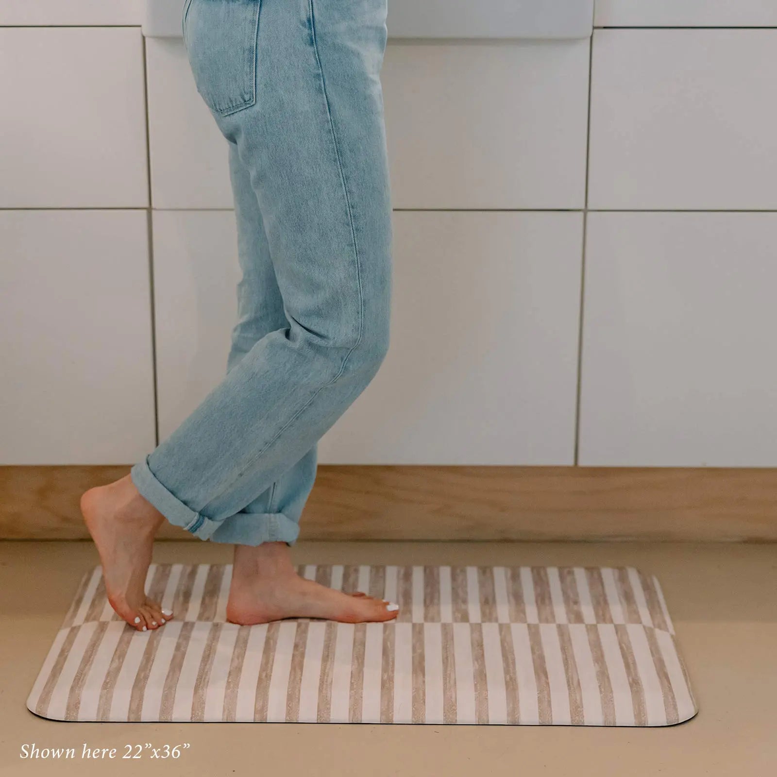 Reese chai beige and white inverted stripe standing mat shown in kitchen in size 22x36 with womans feet standing on the mat