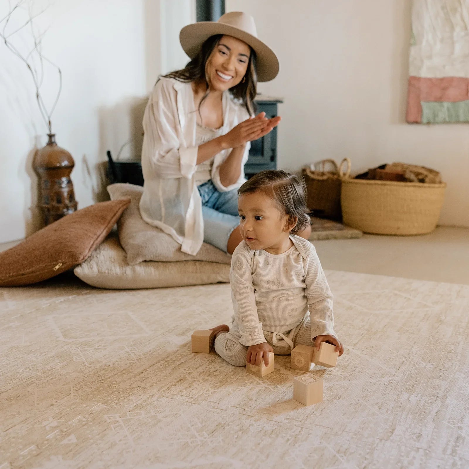 Ula Straw neutral tan minimal boho pattern play mat shown in living room with Mom sitting on pillows and baby boy playing with wooden blocks