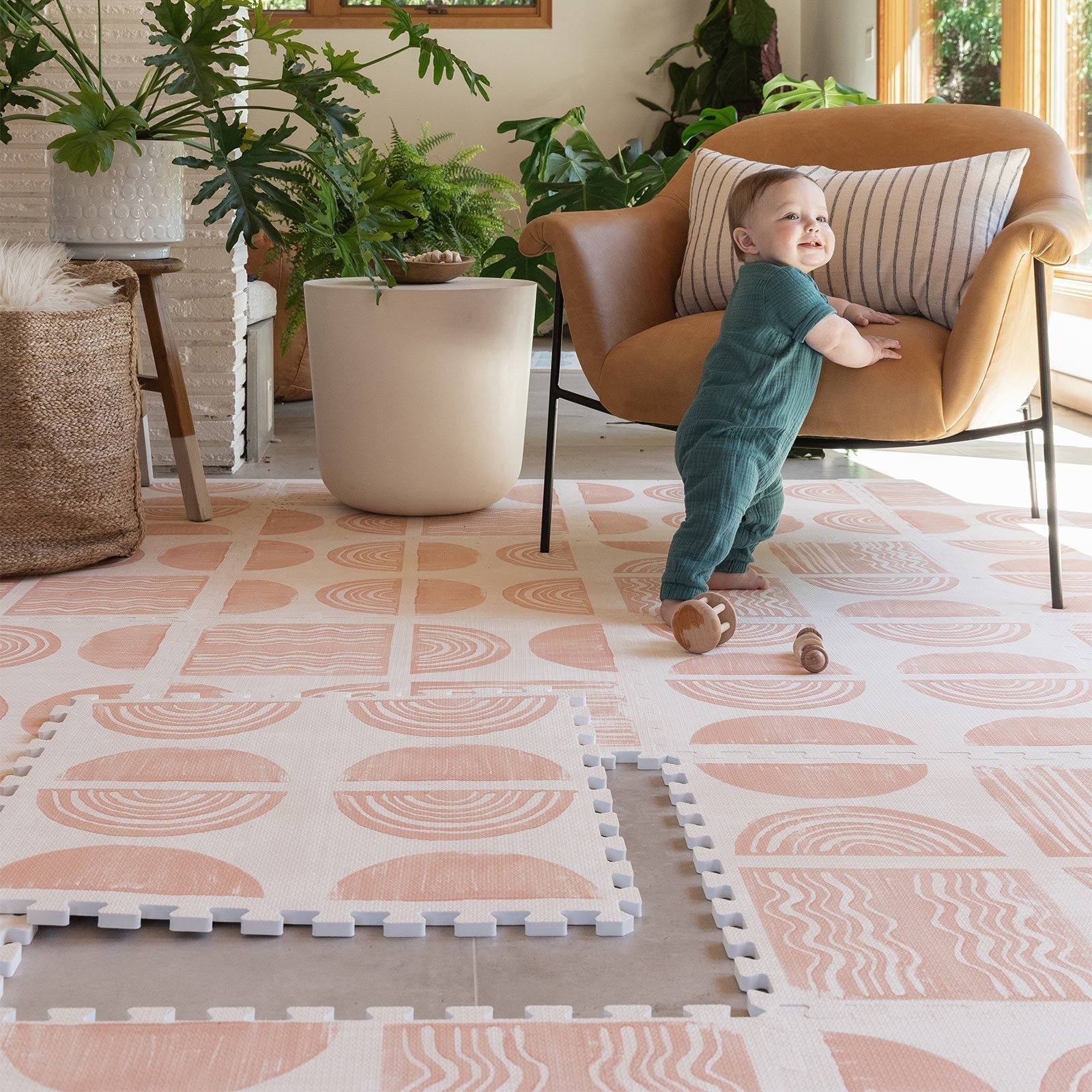 Ada modern minimalist baby play mat in Melon, peach pink and off white. Shown in living room with one tile lifted up and baby standing up against a chair.