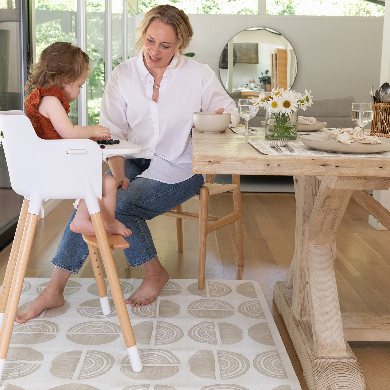 Ada modern minimalist baby high chair mat in Pebble taupe and off white. Shown at kitchen table with Mom feeding baby in high chair.