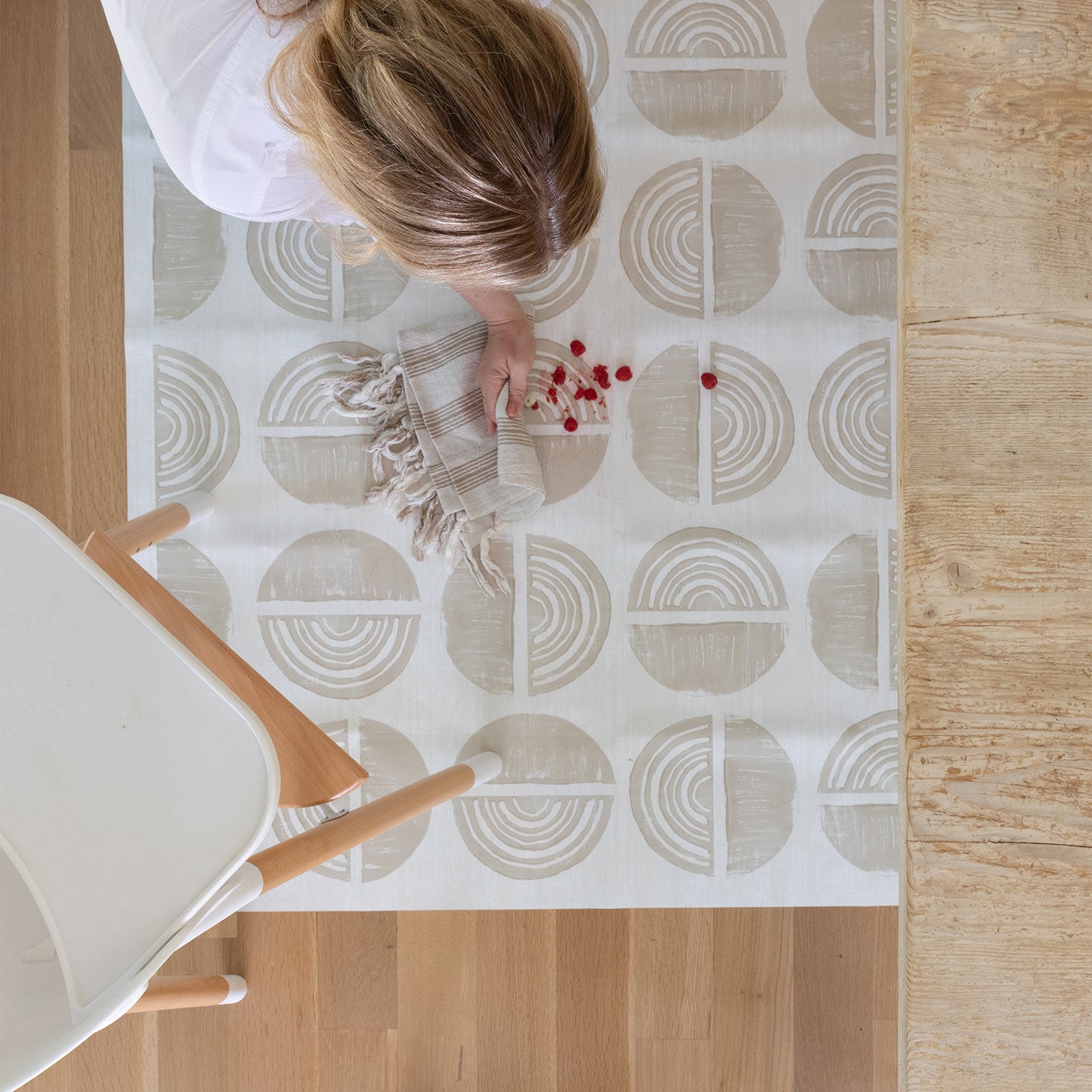 Ada modern minimalist baby high chair mat in Pebble taupe and off white. Shown from above with woman cleaning up a spill under a high chair.