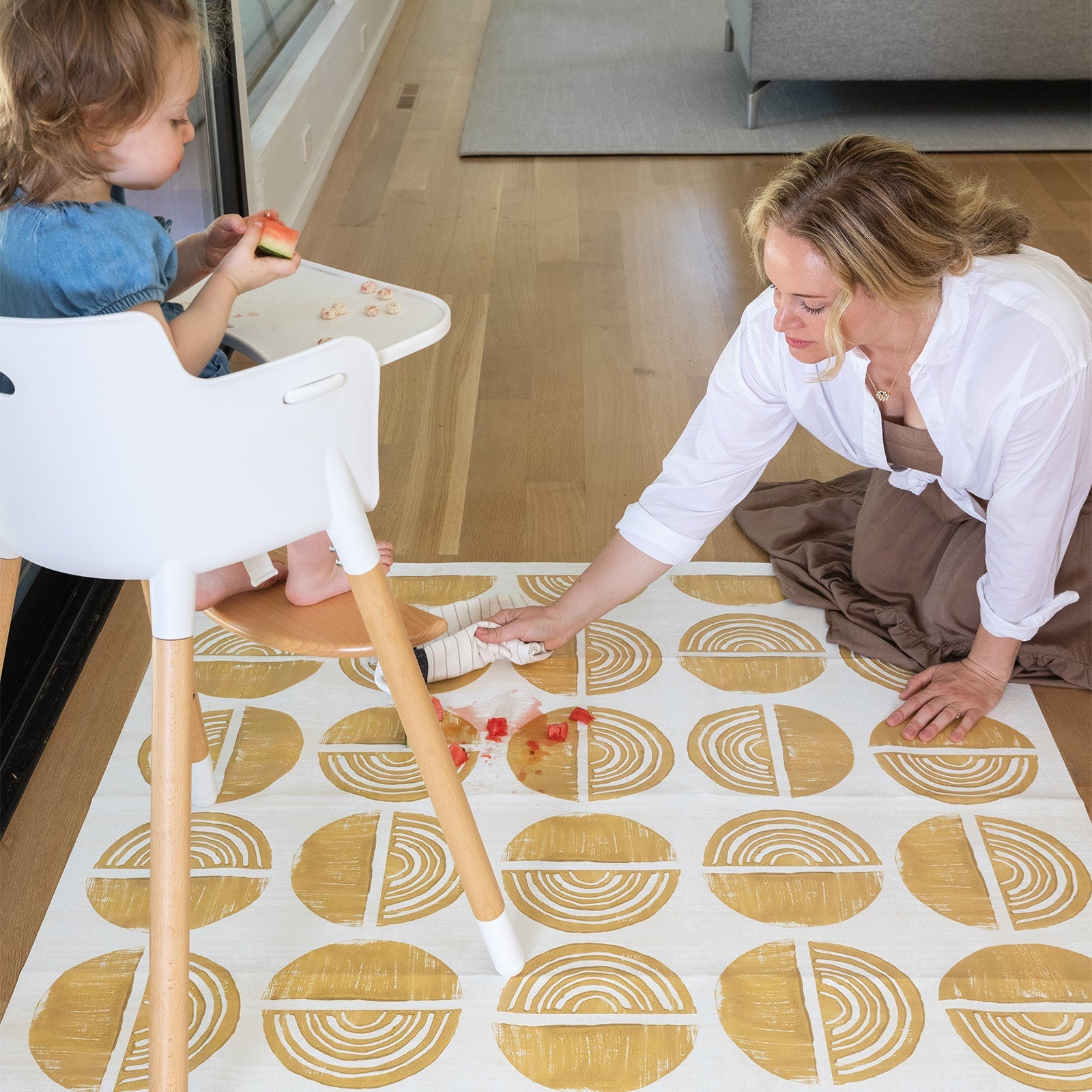 Ada modern minimalist baby high chair mat in Sunflower, mustard yellow and off white. Shown with a baby in a high chair and mom cleaning up a spill.