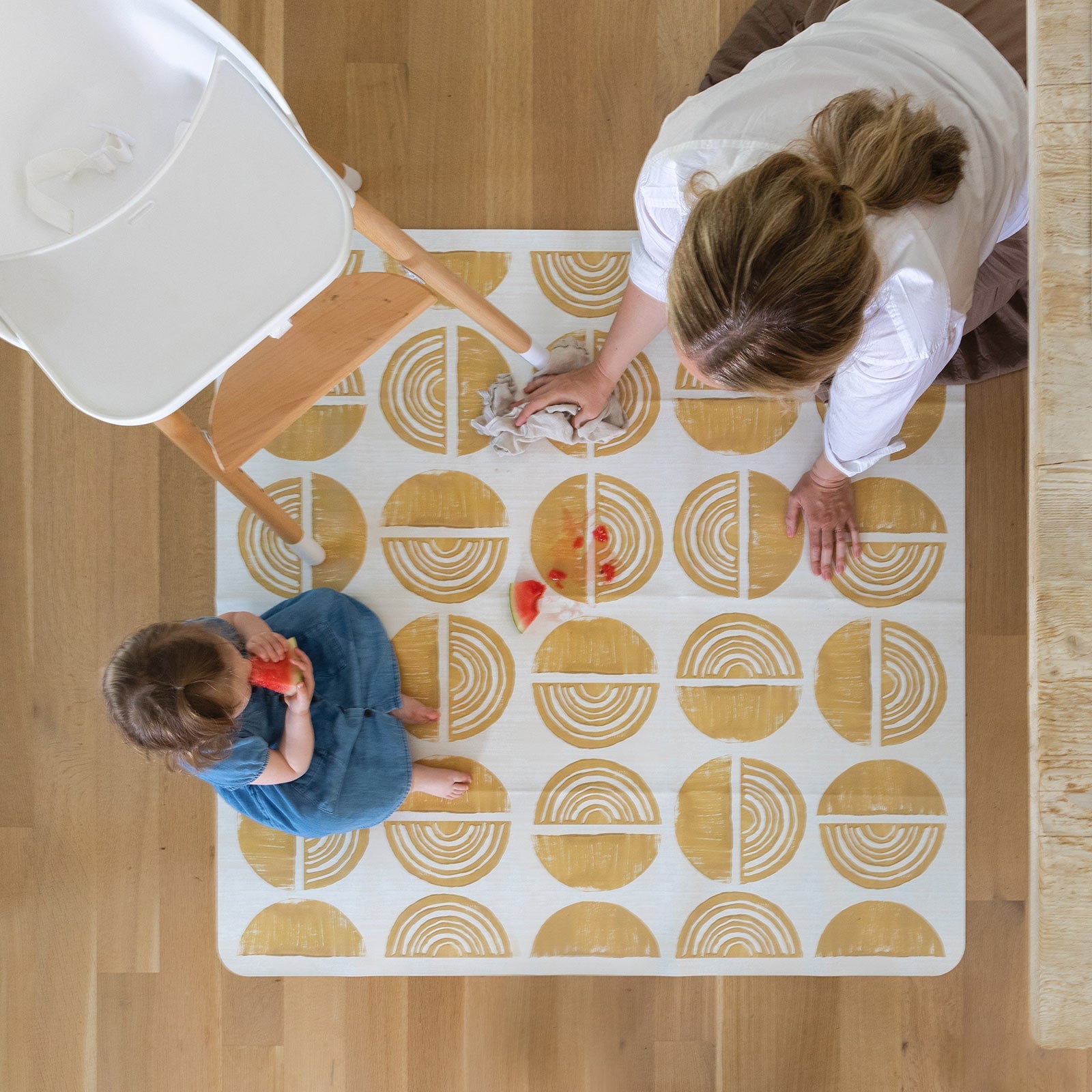 Ada modern minimalist baby high chair mat in Sunflower, mustard yellow and off white. Shot from above with a baby, high chair, and mom cleaning up a spill.