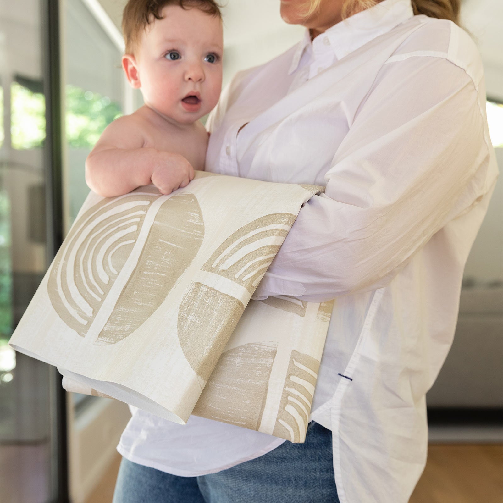 Ada modern minimalist baby high chair mat in Pebble taupe and off white shown folded up being carried with a baby by Mom.