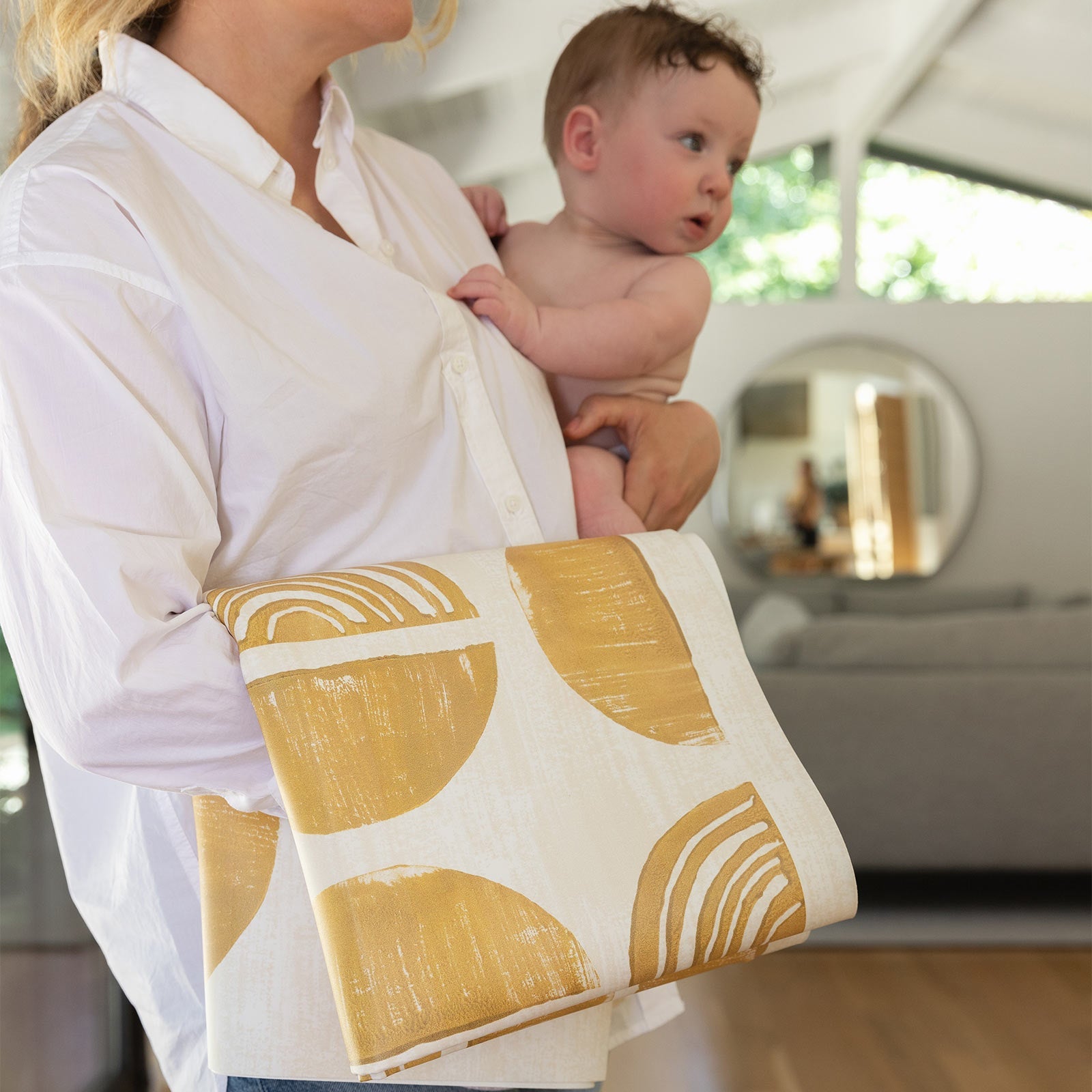 Ada modern minimalist baby high chair mat in Sunflower, mustard yellow and off white folded up being carried with a baby by Mom.